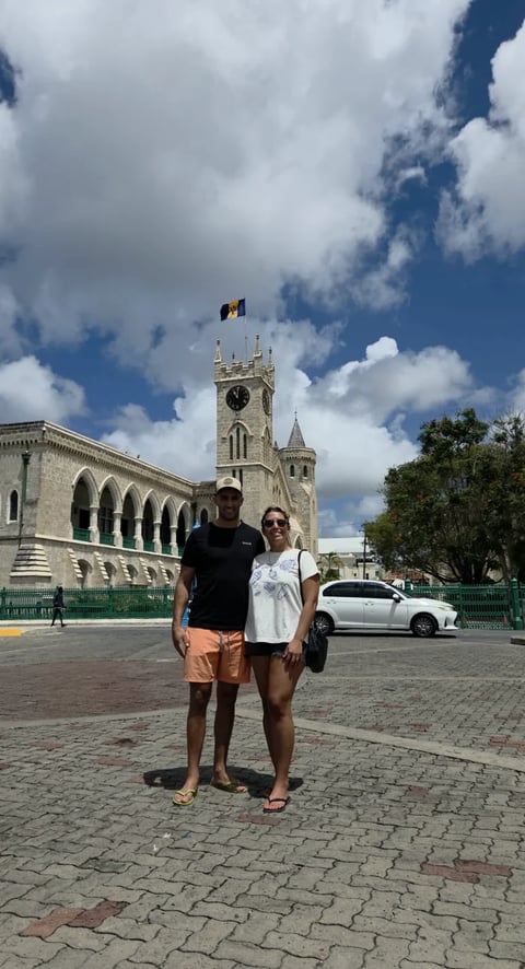 Two people standing in front of a historic stone church with clock tower in Barbados