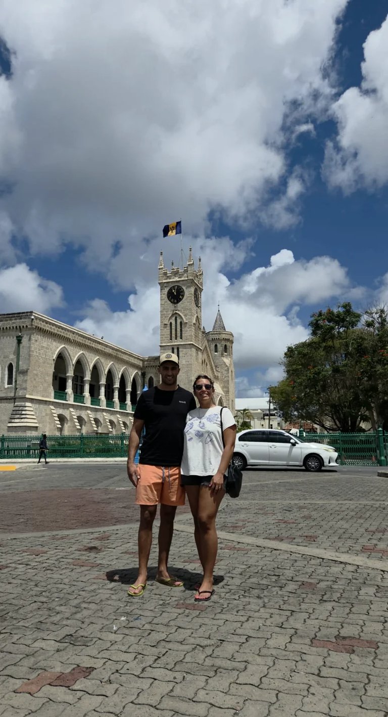 Two people standing in front of a historic stone church with clock tower in Barbados